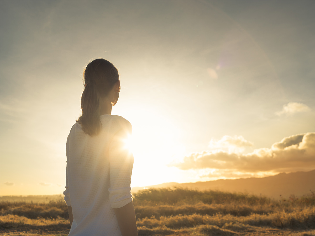 person standing on a field looking at the sun shine behind her