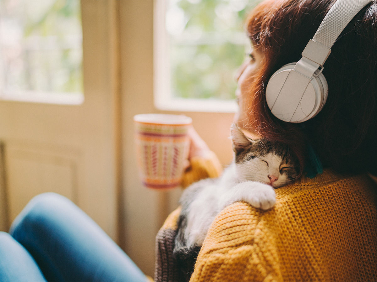 person sitting with headphones on drinking out of a mug and cuddling a cat. looks relaxed